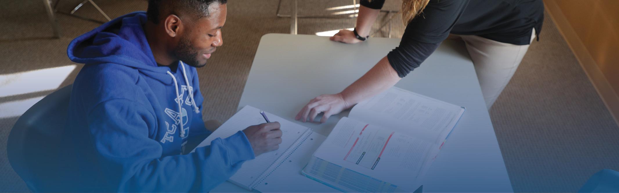 Student at table with open book, writing in notebook with instructor pointing at his page